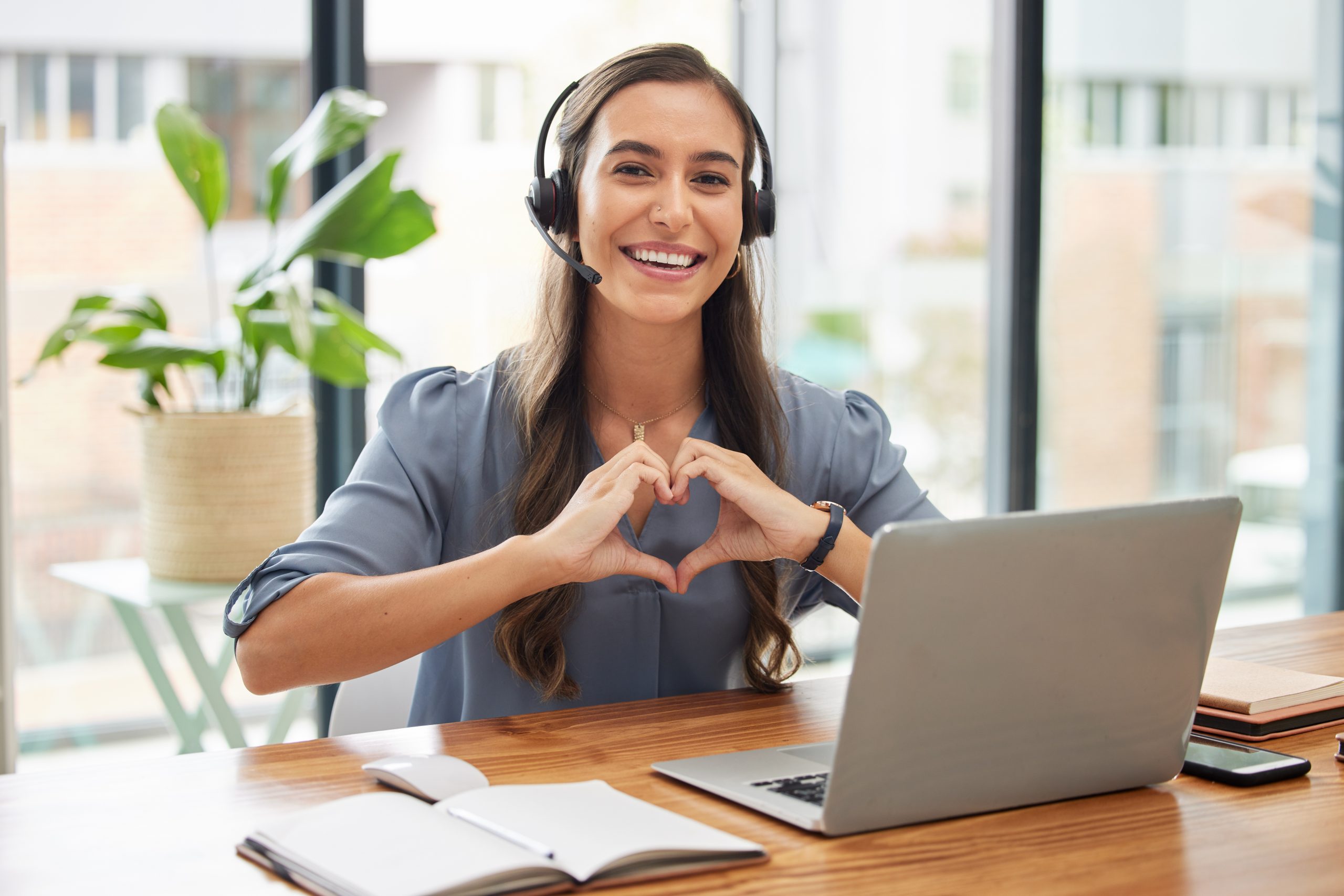 A smiling support professional wearing a headset forms a heart shape with her hands while working at a laptop.