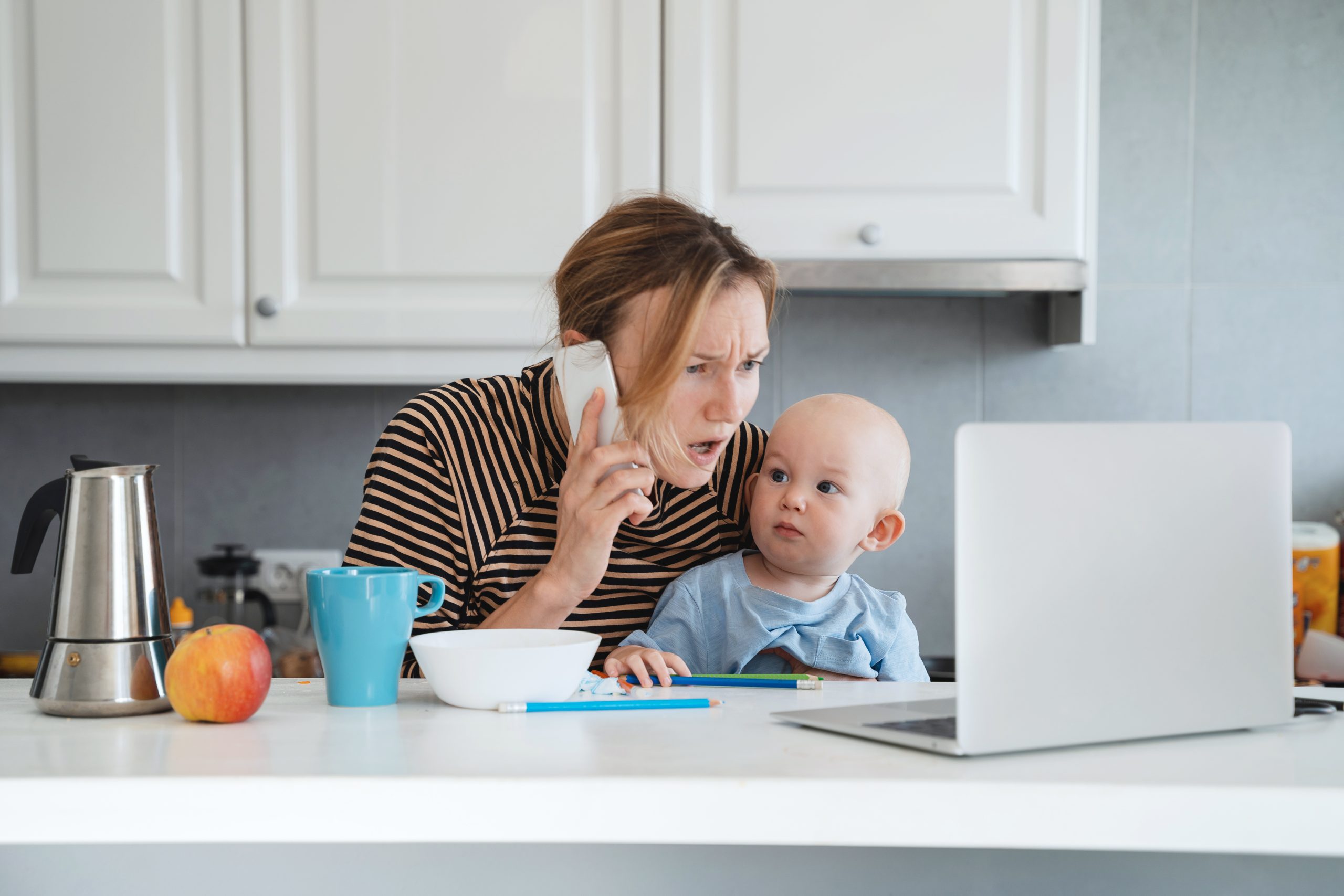 A concerned parent holding a baby speaks on the phone while looking at a laptop in a kitchen setting.
